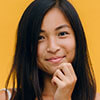 A woman with long dark hair stands in front of a yellow background, looking at the camera and touching her chin with her hand.